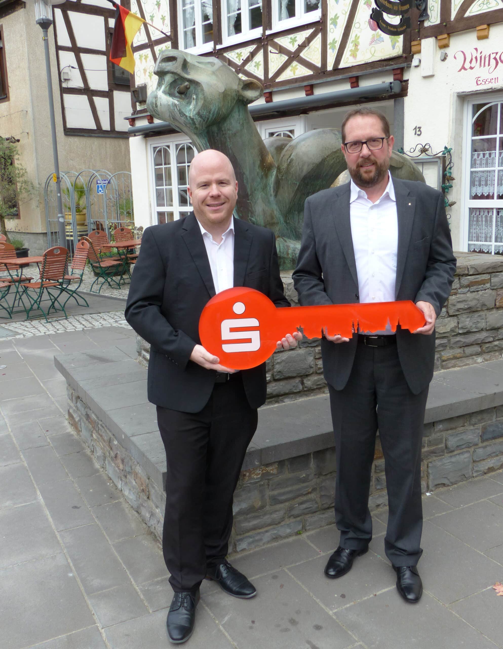 Der neue Filialleiter Patrick Zillich (links) und sein Vorgänger Peter Hammes (rechts) bei der symbolischen Schlüsselübergabe vor dem Tatzelwurmbrunnen auf dem Marktplatz von Kobern-Gondorf. Foto: Manfred Gerz.