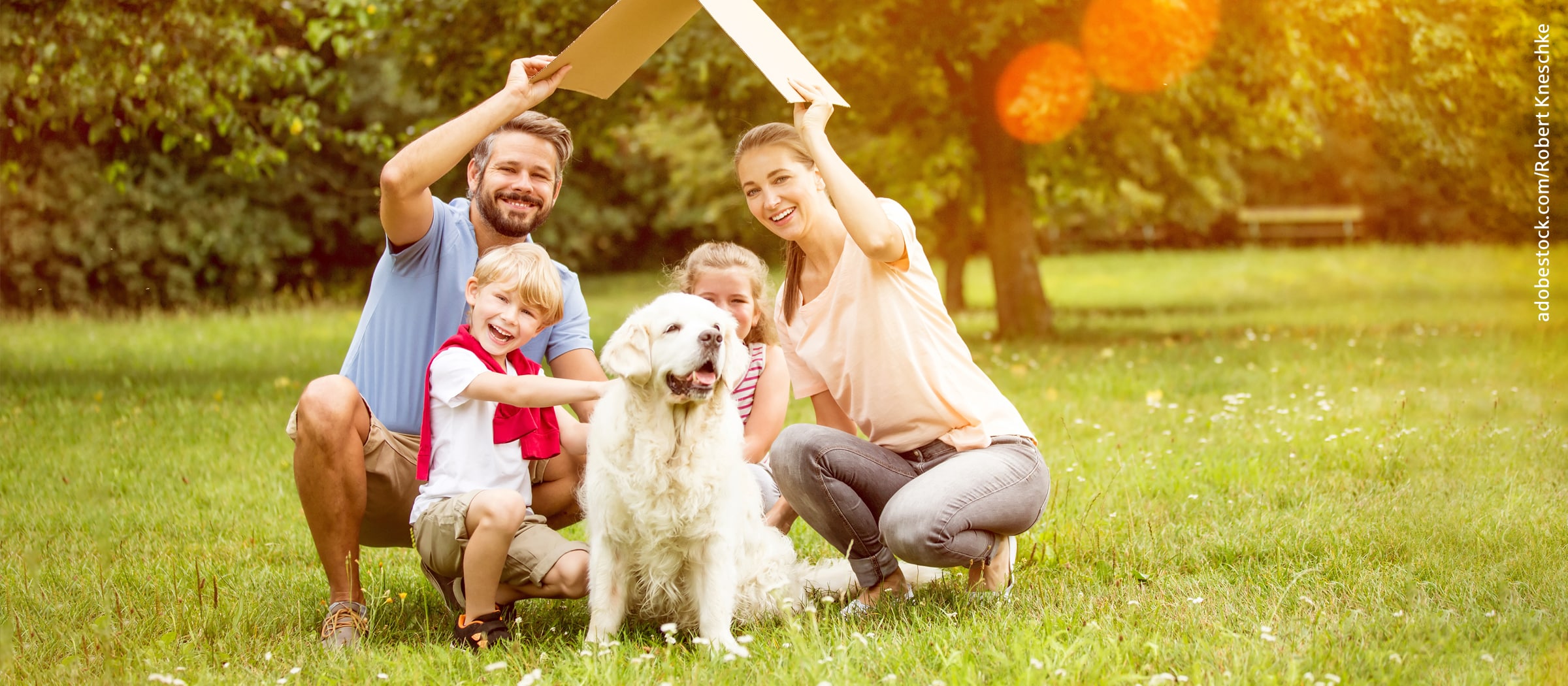 Eine Familie sitzt zusammen mit einem Hund auf einer Wiese und hält ein Papp-Papier, gefaltet zu einem Dach, über sich.
