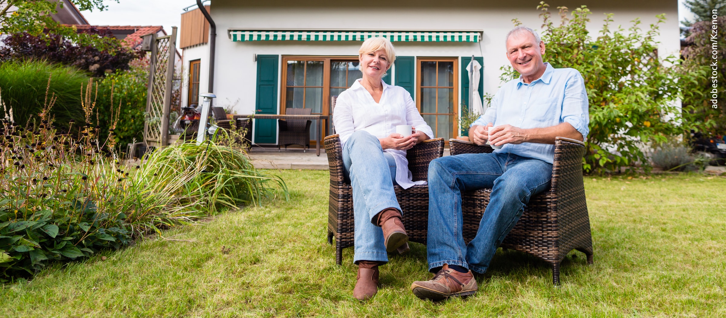 Zwei Senioren sitzen im Garten - im Hintergrund ihr Haus mit Terasse